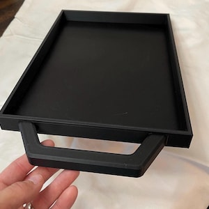 May include: A black rectangular tray with a handle. The tray is made of a solid material and has a raised edge. The handle is a thick, rectangular shape. The tray is shown on a white surface.