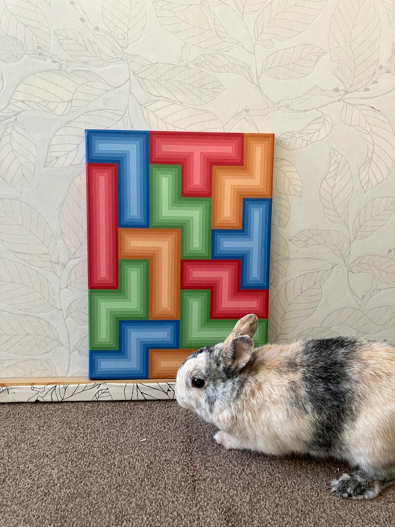 May include: A colorful abstract painting with a geometric design in shades of red, blue, green, and orange. A small rabbit with black and white fur is in the foreground. The painting is on a rectangular canvas.