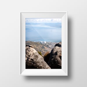 May include: A panoramic view from a mountaintop overlooking a city and the ocean. The city is in the foreground, with a large stadium visible. The ocean is in the background, with a blue sky and white clouds.