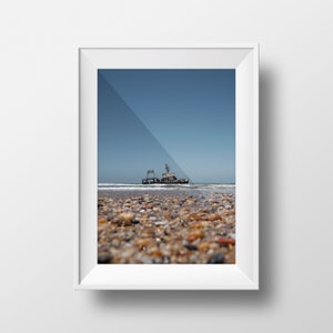 Peut inclure: Une photographie encadrée en blanc d'un navire rouillé échoué sur une plage avec un ciel bleu en arrière-plan.