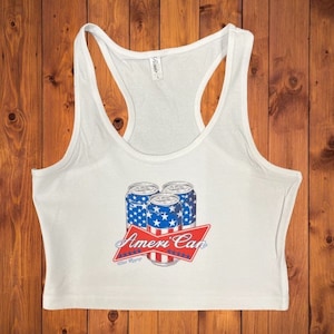 May include: White tank top with a cropped hem featuring a graphic of three American flag-themed beer cans with the text "Ameri'Can".