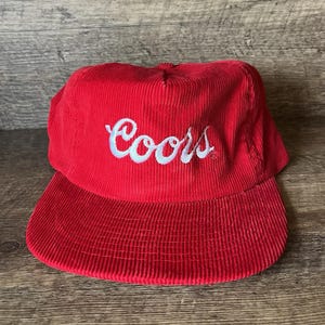 Peut inclure: Casquette de baseball rouge en velours côtelé avec une visière incurvée. Le mot "Coors" est brodé en blanc sur le devant. Un chapeau de style classique, adapté aux tenues décontractées.