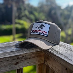 Op de afbeelding: Een bruinbeige baseballpet met een platte klep en een touwdetail. De pet heeft een rechthoekige patch met de tekst "Winchester Rifles and Shotguns" in rood en wit, en een afbeelding van een persoon te paard.