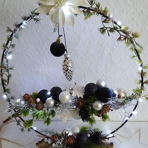 Könnte beinhalten: Ein kreisförmiger Weihnachtskranz mit schwarzem Metallrahmen, geschmückt mit Grün, Tannenzapfen und verschiedenen Ornamenten in Schwarz, Silber und Weiß. Der Kranz ist mit kleinen weißen Lichtern beleuchtet und hat oben eine große weiße Blume.