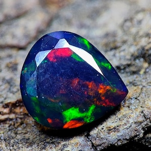 Può includere: Una gemma di opale nero sfaccettata a forma di goccia con vibranti colori interni rossi, verdi e arancioni. La gemma è posta su una superficie di pietra strutturata, marrone e grigia, che ne evidenzia la brillantezza.