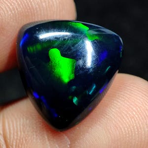 Puede incluir: Una piedra preciosa de ópalo negro triangular pulido, con vibrantes destellos verdes y azules. La gema se sostiene entre los dedos, mostrando su juego de colores iridiscente. La superficie del ópalo es lisa y reflectante, resaltando su belleza natural.