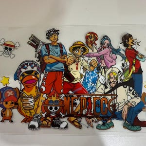 May include: A clear plastic sticker featuring a colorful illustration of characters from the anime series "One Piece." The design includes various characters in action poses, with the series' logo prominently displayed in a brown, metallic font.