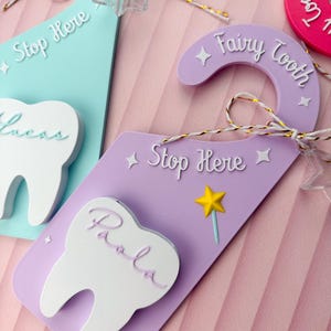May include: Close-up of several tooth-shaped door hangers in pastel colors. The purple hanger reads "Fairy Tooth" and "Stop Here." A white tooth shape with the name "Paola" is attached. Other hangers are light blue and pink.