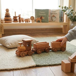 May include: A wooden toy train with two cars, the engine is engraved with the name "Brian". The train is on a green and white rug. Other wooden toys, including animal figures and a stacking tower, are on a shelf in the background.