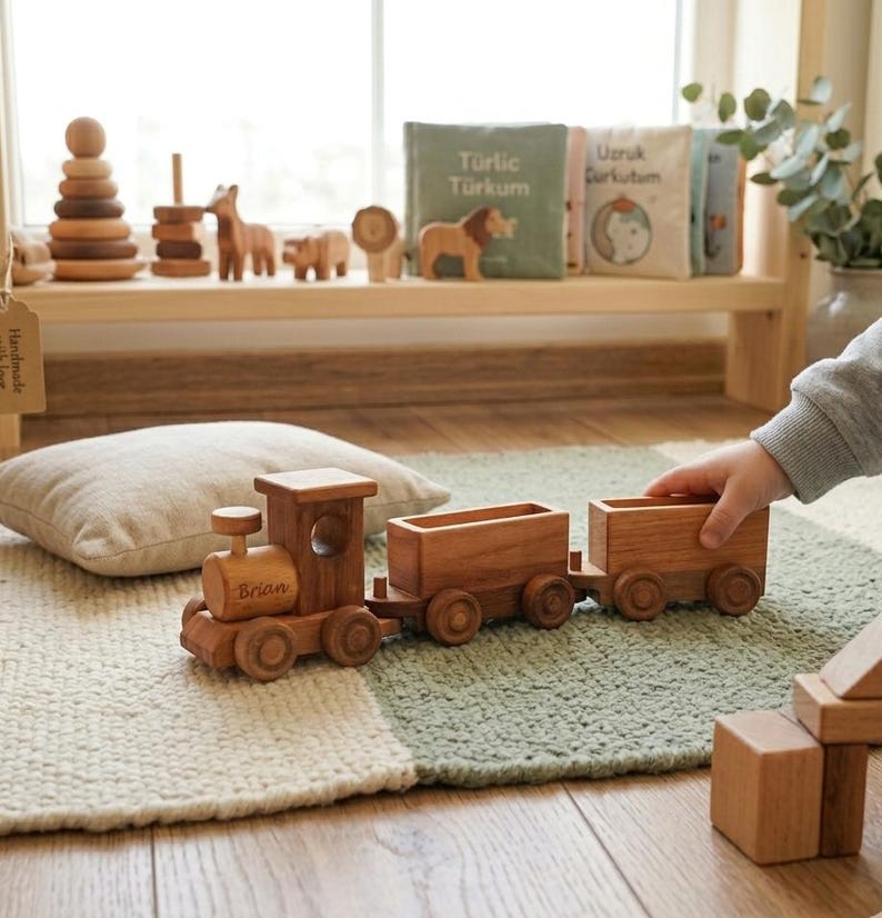 May include: A wooden toy train with two cars, the engine is engraved with the name "Brian". The train is on a green and white rug. Other wooden toys, including animal figures and a stacking tower, are on a shelf in the background.