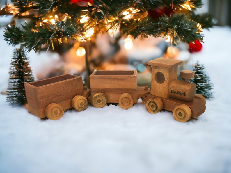 May include: A wooden toy train set with three cars, including an engine with the name "Rafael" engraved on the side. The train is brown and sits on a snowy surface, with a Christmas tree and lights in the background.
