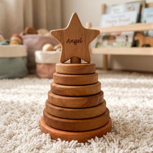 May include: A wooden stacking toy with a star topper. The star is engraved with the name "Angel". The toy has a series of graduated wooden rings. The toy is made of light brown wood and sits on a white rug.