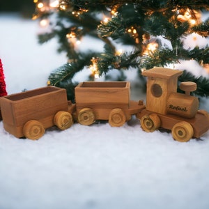 May include: A wooden toy train with three cars, including an engine with the name "Rafael" engraved on the side. The train is brown and sits on a snowy surface, with a small red Christmas tree and a decorated Christmas tree in the background.