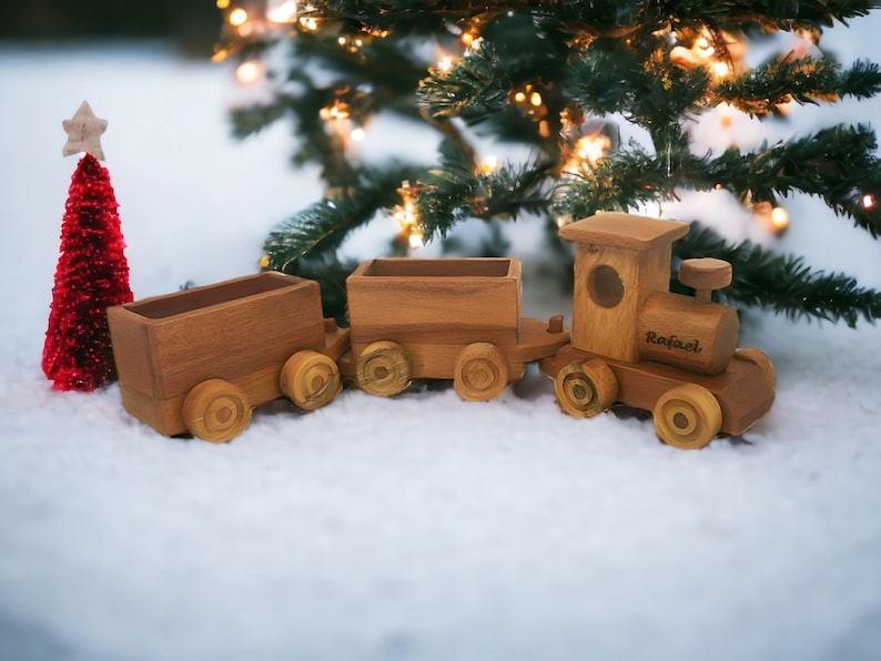 May include: A wooden toy train with three cars, including an engine with the name "Rafael" engraved on the side. The train is brown and sits on a snowy surface, with a small red Christmas tree and a decorated Christmas tree in the background.
