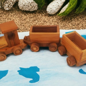 May include: A wooden toy train set with three cars. The engine has the name "Brian" engraved on it. The train is made of light brown wood and has round wheels. The train is on a blue and white surface.