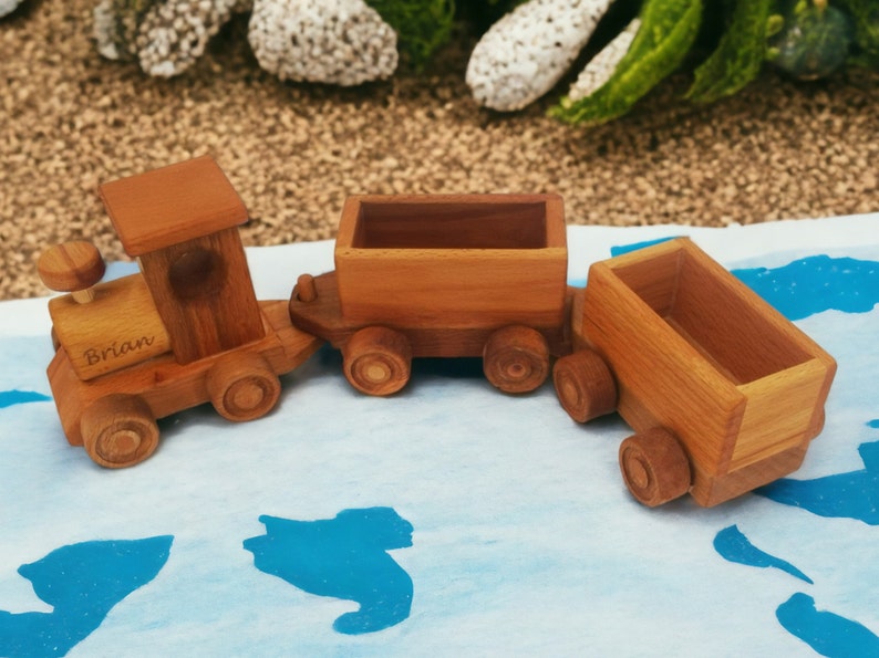 May include: A wooden toy train set with three cars. The engine has the name "Brian" engraved on it. The train is made of light brown wood and has round wheels. The train is on a blue and white surface.