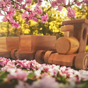 May include: A wooden toy train with two attached cars, set against a blurred background of pink flowers and a green and yellow landscape. The train is made of light brown wood and has the name "Brian" engraved on the side.
