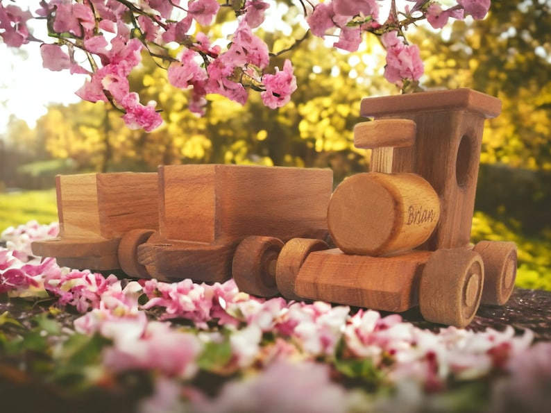 May include: A wooden toy train with two attached cars, set against a blurred background of pink flowers and a green and yellow landscape. The train is made of light brown wood and has the name "Brian" engraved on the side.
