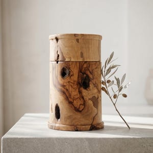 May include: A cylindrical wooden container with a lid, featuring a natural wood grain pattern and several knot holes. The container is light brown and sits on a light-colored surface. A sprig of olive branches rests beside it.