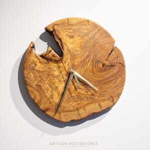 May include: A round wall clock crafted from a natural olive wood slab with a live edge, featuring prominent wood grain patterns, a dark knot, and three slender gold-toned metal hands. The text ARTISAN WOODWORKS is printed at the bottom.