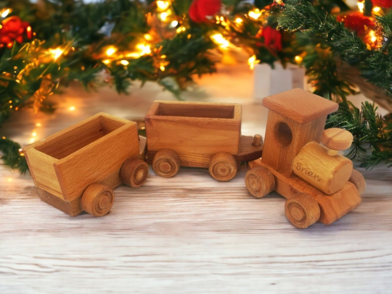 May include: A wooden toy train with three cars, including an engine and two cargo cars. The train is made of light brown wood and has wheels. The engine has the name "Brian" engraved on it. The background includes Christmas decorations.