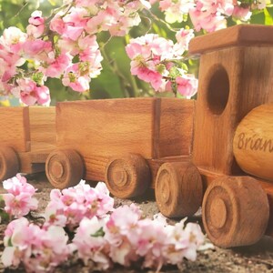 May include: A wooden toy train with two cars, featuring a natural wood finish. The train has round wheels and a cylindrical smokestack. The name "Brian" is engraved on the side. Pink flowers are in the background.