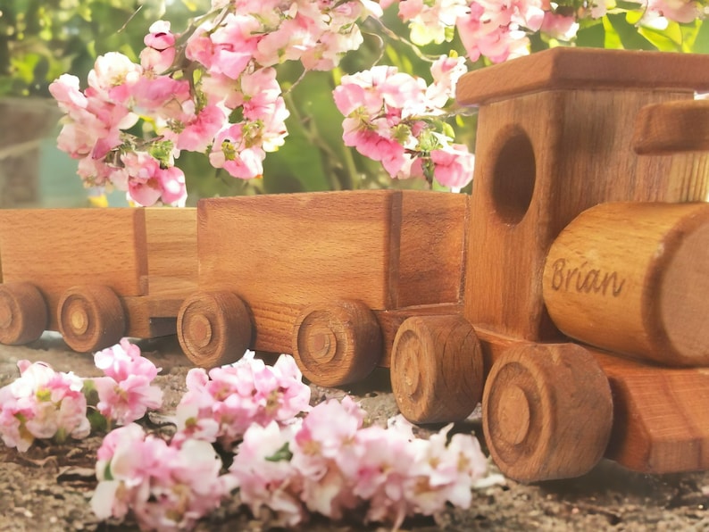 May include: A wooden toy train with two cars, featuring a natural wood finish. The train has round wheels and a cylindrical smokestack. The name "Brian" is engraved on the side. Pink flowers are in the background.