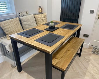 May include: A wooden dining table with black metal legs and a matching wooden bench. The table has four place settings with black placemats and a small potted plant in the centre.