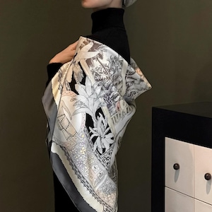 May include: A large square silk scarf featuring a detailed black, white, and grey illustrative print of tropical foliage, architectural elements, and a central crest. The scarf has a wide dark grey border and is draped over a black turtleneck jumper.