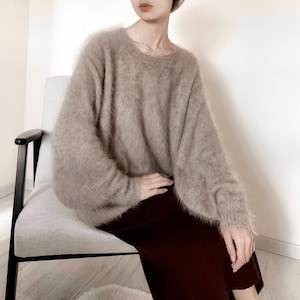 May include: A beige, oversized sweater with long sleeves. The sweater has a soft, fuzzy texture and is paired with a dark burgundy skirt. The person is seated in a light gray chair with black armrests.
