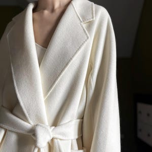 Könnte beinhalten: Ein langärmeliger, cremeweißer Wickelmantel mit breitem Reverskragen und einem passenden Bindegürtel in der Taille. Das Kleidungsstück ist aus einem weichen, gewalkten Material gefertigt und besticht durch ein schlichtes Design ohne sichtbare Knöpfe.