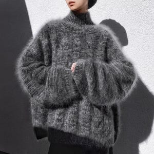Op de afbeelding: Een grijze, oversized trui met een hoge hals en lange, uitlopende mouwen. De trui heeft een getextureerde, fuzzy uitstraling en is gemaakt van een zacht materiaal. Dit kledingstuk is een stijlvol kledingstuk voor koeler weer.