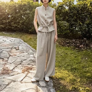 Op de afbeelding: Een beige mouwloze top met een opstaande kraag, een knoopsluiting aan de voorzijde en een taille met trekkoord, gecombineerd met een bijpassende broek met wijde pijpen. De outfit wordt afgemaakt met witte instappers voor een gecoördineerde set.