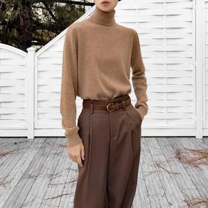 Può includere: Un dolcevita beige abbinato a pantaloni a pieghe marroni e una cintura in pelle marrone con fibbia dorata. L'outfit ha una palette di colori neutri, creando un look classico e versatile.