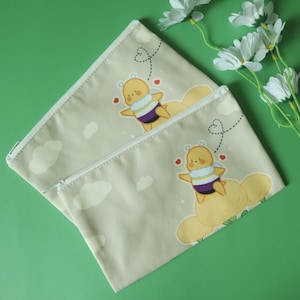 May include: Two beige canvas zipper pouches with a cartoon bee design. The bee has a purple and white scarf and is riding a yellow cloud. White clouds and hearts are also printed on the pouches.