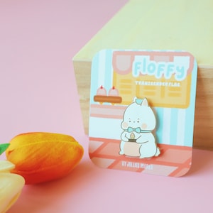 Op de afbeelding: Een gouden emaille pin met een wit cartoonfiguur dat een ijsje vasthoudt. Het figuur draagt een blauwe strik en een blauwe hoed. De pin is op een kaart met de tekst "Floffy Transgender Flag" en "By Jellies World".