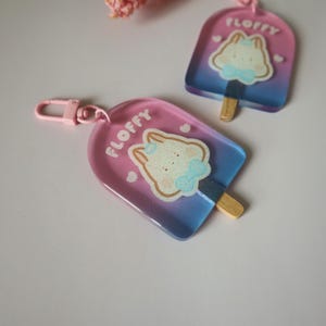 May include: Two popsicle-shaped keychains with a pink and blue gradient. Each keychain features a cartoon bunny with a blue bow tie and the word "FLOFFY". One keychain has a pink clasp.