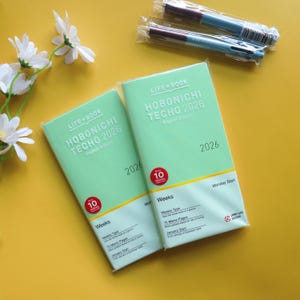 Puede incluir: Dos agendas Hobonichi Techo 2026 en verde menta, edición en inglés, con el texto "LIFE BOOK" y "HOBONICHI TECHO 2026" impreso en la portada. Dos bolígrafos en embalaje plástico. Sobre una superficie amarilla.
