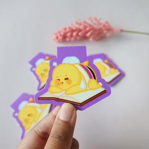 May include: A collection of purple and yellow bee-themed magnetic bookmarks. Each bookmark features a cartoon bee resting on a book. The bee is yellow with white, purple, and black stripes. The background is white with a pink flower.