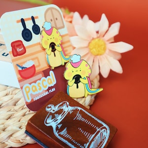 May include: Two enamel pins featuring a yellow cartoon character wearing a chef's hat and holding a black pan. The character is labeled "Pascal" and "Pansexual Flag". The pins are on a brown background with a white jar illustration.