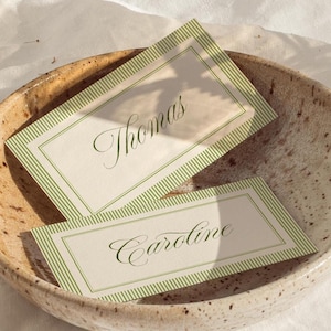 Könnte beinhalten: Eine gesprenkelte Keramikschale enthält zwei Namenskarten. Die Karten haben einen weißen Hintergrund mit grün gestreiften Rändern und elegante Schriftnamen: "Thomas" und "Caroline". Die Schale steht auf einer weißen, strukturierten Oberfläche.