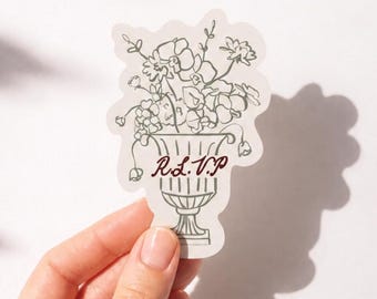 Könnte beinhalten: Ein gestanzter Aufkleber mit einer schwarzen Strichzeichnung eines Blumenarrangements in einer klassischen Urne. Der Text R.S.V.P. ist in einer dunkelroten Schreibschrift in der Mitte der Urne auf einem cremeweißen Hintergrund gedruckt.