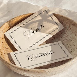 Puede incluir: Un cuenco de cerámica moteada contiene dos tarjetas rectangulares. Las tarjetas tienen un fondo crema con un borde negro y una escritura elegante. Una tarjeta dice "Thomas" y la otra "Candela".