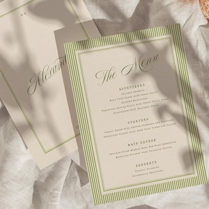 May include: Two beige menu cards with green striped borders and elegant script. One card reads "The Menu" and lists appetizers, starters, main course, and desserts. A half-eaten orange and a beige mesh bag rest on a white linen surface.