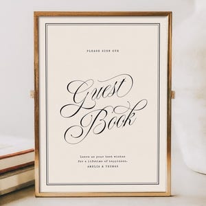 May include: A rectangular gold-toned metal frame containing a cream-colored sign with black text. The sign reads: PLEASE SIGN OUR Guest Book. Leave us your best wishes for a lifetime of happiness. AMELIA & THOMAS. The frame has a thin double-line border.