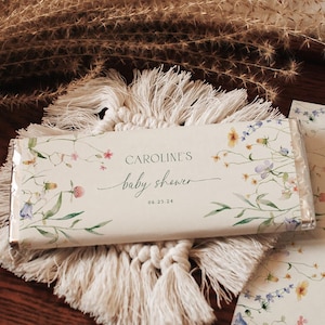 May include: A white chocolate bar with a floral design and the text "CAROLINE'S baby shower 06.27.24" on a wooden tray with a white tassel.