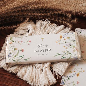 May include: A white chocolate bar with a floral design and the text "Olivia's Baptism 06.25.21". The chocolate bar is on a white crocheted mat with tassels.