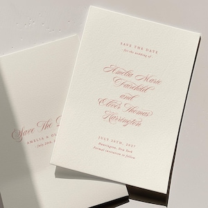 May include: Two ivory wedding save-the-date cards with burgundy script. The cards read "Save the Date" and list the names Amelia Marie Fairchild and Oliver Thomas Harrington, with the date July 20th, 2027.