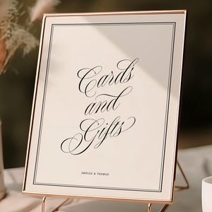 Op de afbeelding: Een rechthoekig bord in een dun roségouden frame met de zwarte tekst "Cards and Gifts" en "AMELIA & THOMAS" in een schreeflettertype. Het bord staat op een roségouden draadstandaard, vergezeld door een witte vaas met droogbloemen en een wit kopje.