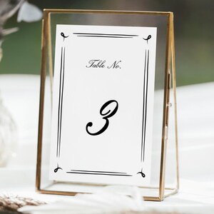 May include: A gold-framed table number card with the number 3 in a decorative font. The card reads "Table No." at the top. The frame is rectangular and made of a thin, gold metal. The background is blurred, suggesting an outdoor setting.